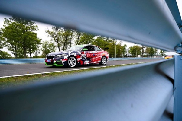Race car number 62 with red, white, and black livery on a racetrack, viewed through blue safety barriers with trees in the background